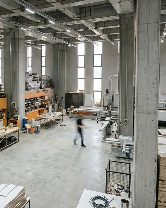 Gösteri Woodworks 5.000 m² entegre üretim tesisinde çalışan operatör; arka planda ahşap işleme makineleri ve yüksek tavanlı atölye alanı.
